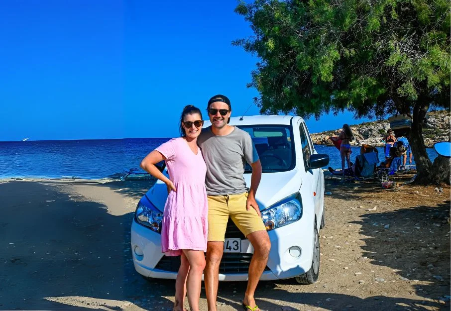 A couple standing in front of a hire car at a beach in Europe